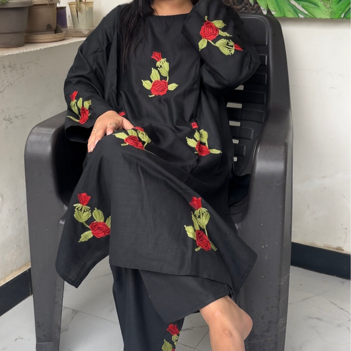 Person wearing a black outfit with red floral embroidery sitting on a chair.